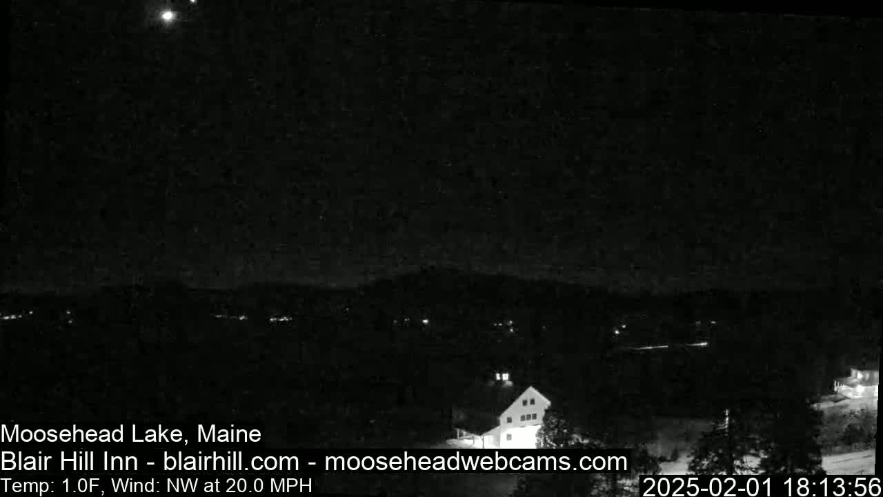 A nighttime, long shot shows a dark landscape with a few scattered lights under a mostly clear sky with a northwest wind blowing at 20 mph and a temperature of 1 degree Fahrenheit.