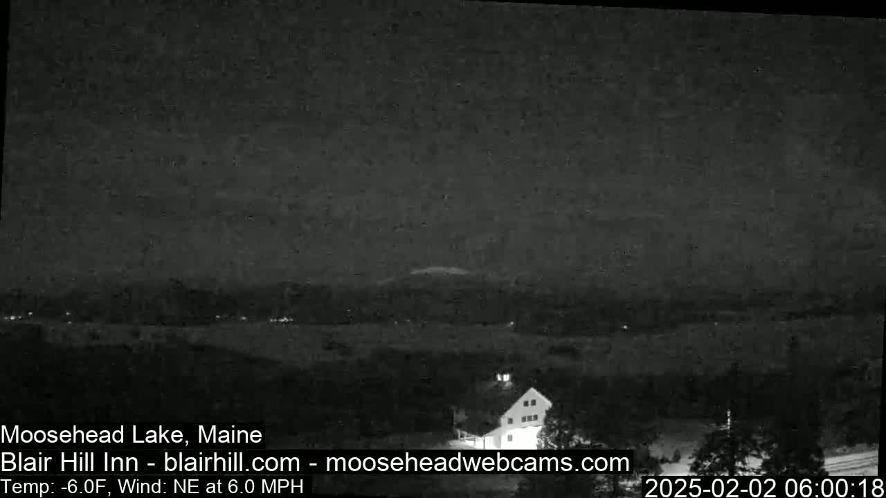 A nighttime, low-light view of a lake and distant lights with a building in the foreground, under a clear sky with a temperature of -6.0°F and a northeasterly wind of 6.0 mph.