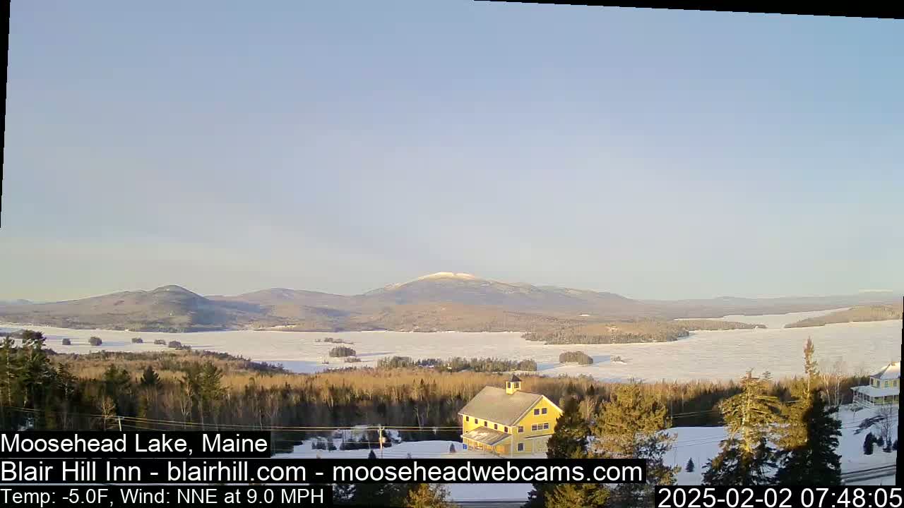 A partly frozen lake stretches to snow-covered mountains under a clear, light blue sky, with a yellow building and evergreen trees in the foreground.