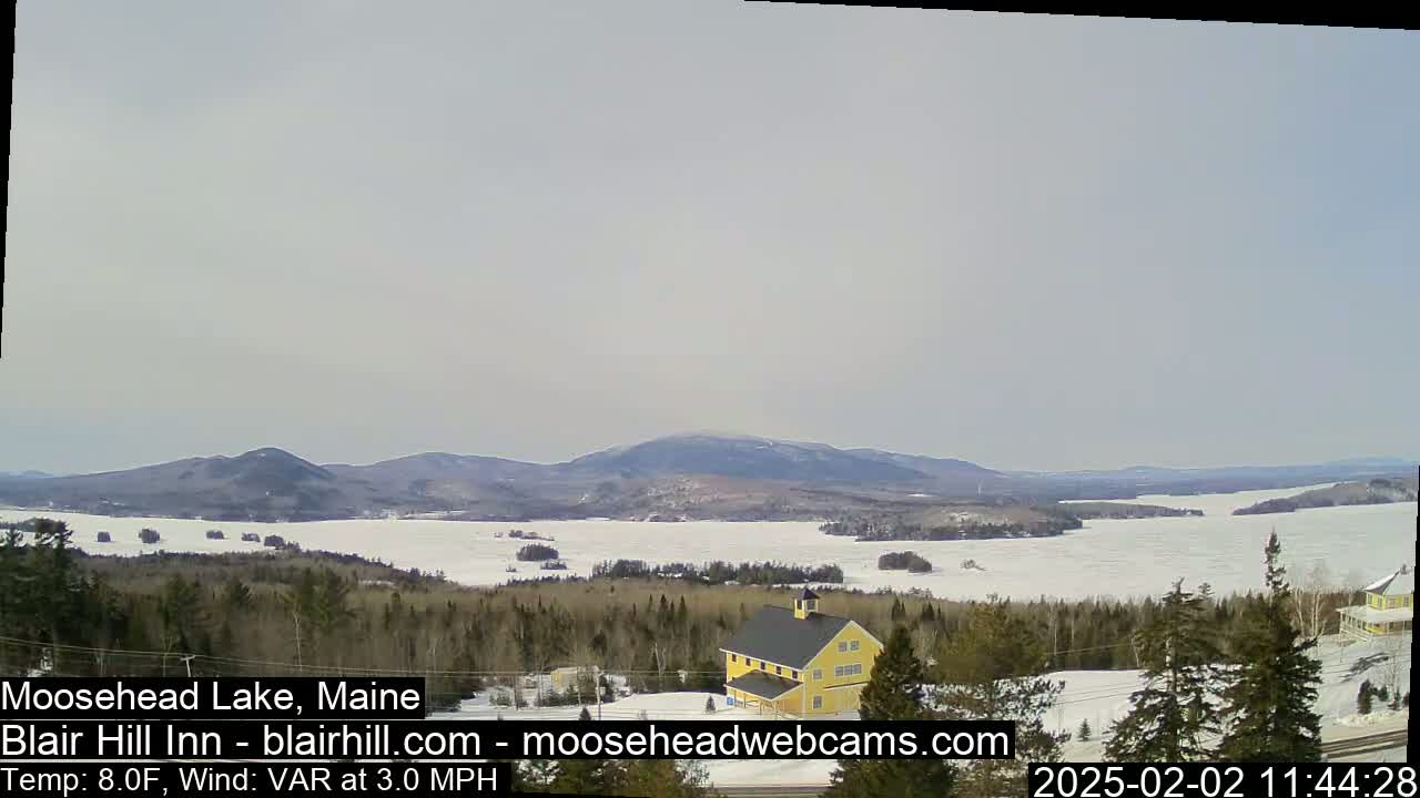 A partly frozen lake is viewed from a hilltop, with a yellow building and a forest in the foreground and distant mountains under a pale sky.