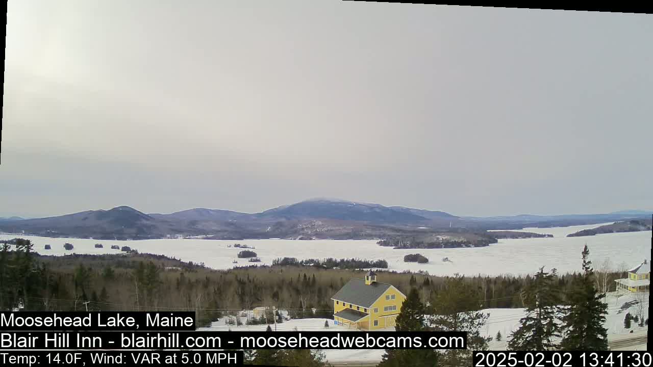 A partly frozen lake is visible from a snowy hilltop, with a yellow building in the foreground and distant mountains under a cloudy sky.