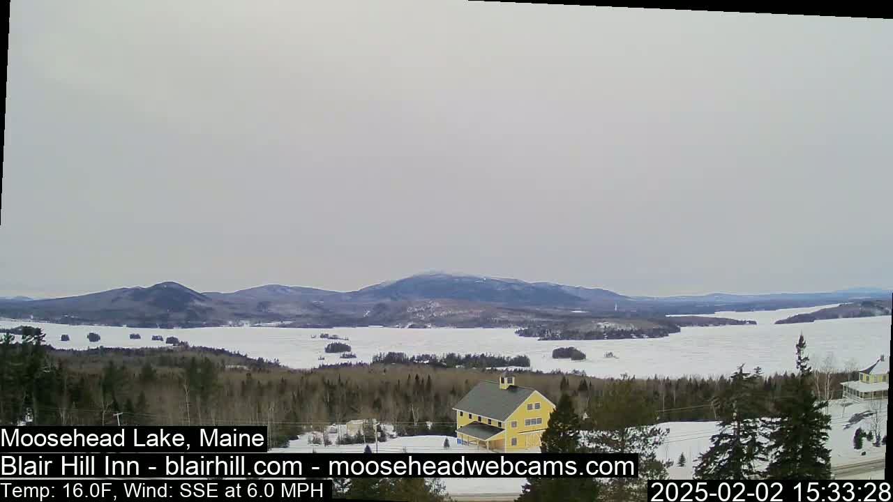 A partially frozen lake is visible from a high vantage point, with a yellow building and snow-covered terrain in the foreground under a cloudy sky.