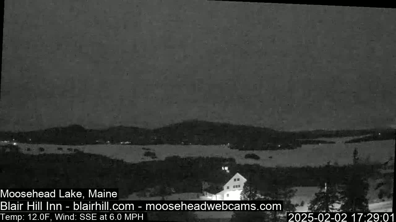 A nighttime view of a snow-covered landscape with a lake and distant hills under a dark, clear sky, with a light wind from the south-southeast.