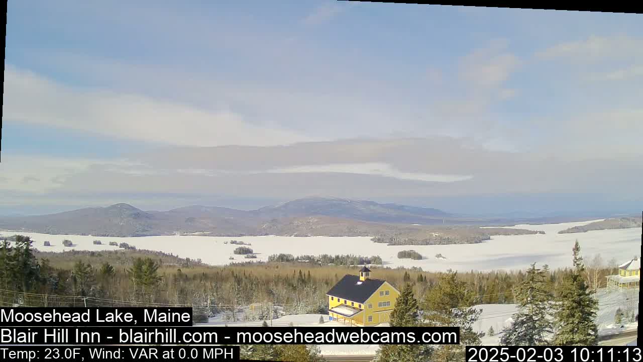 A partly cloudy day reveals a snow-covered lake and distant mountains, with a yellow building situated in the foreground amidst evergreen trees.