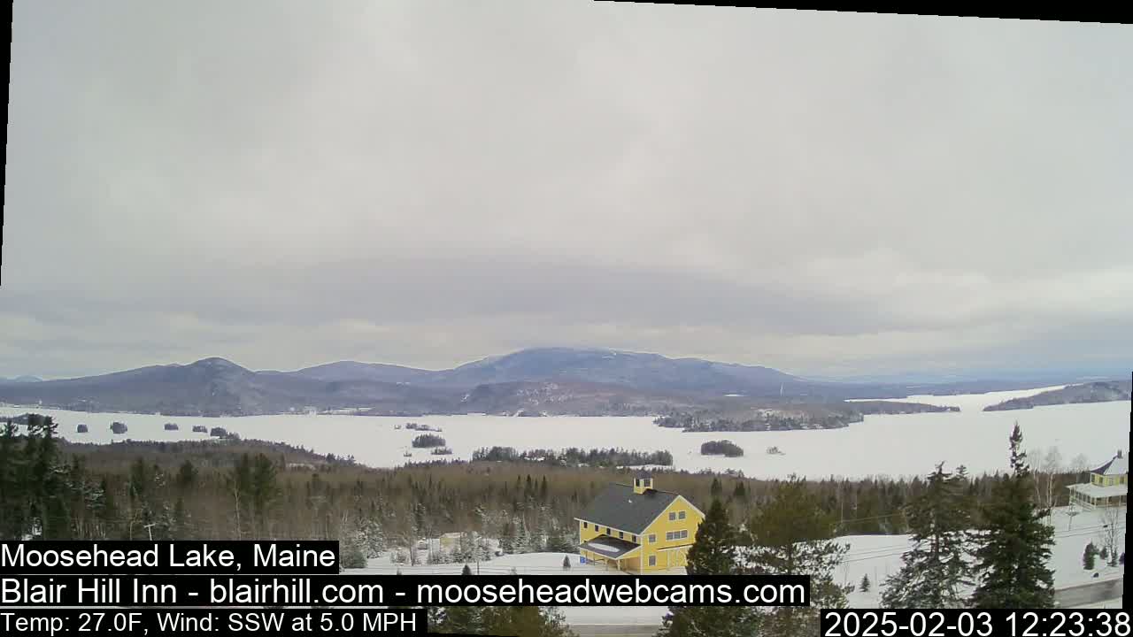 A snow-covered landscape overlooks a frozen lake and distant mountains under an overcast sky, with a yellow building visible in the foreground.