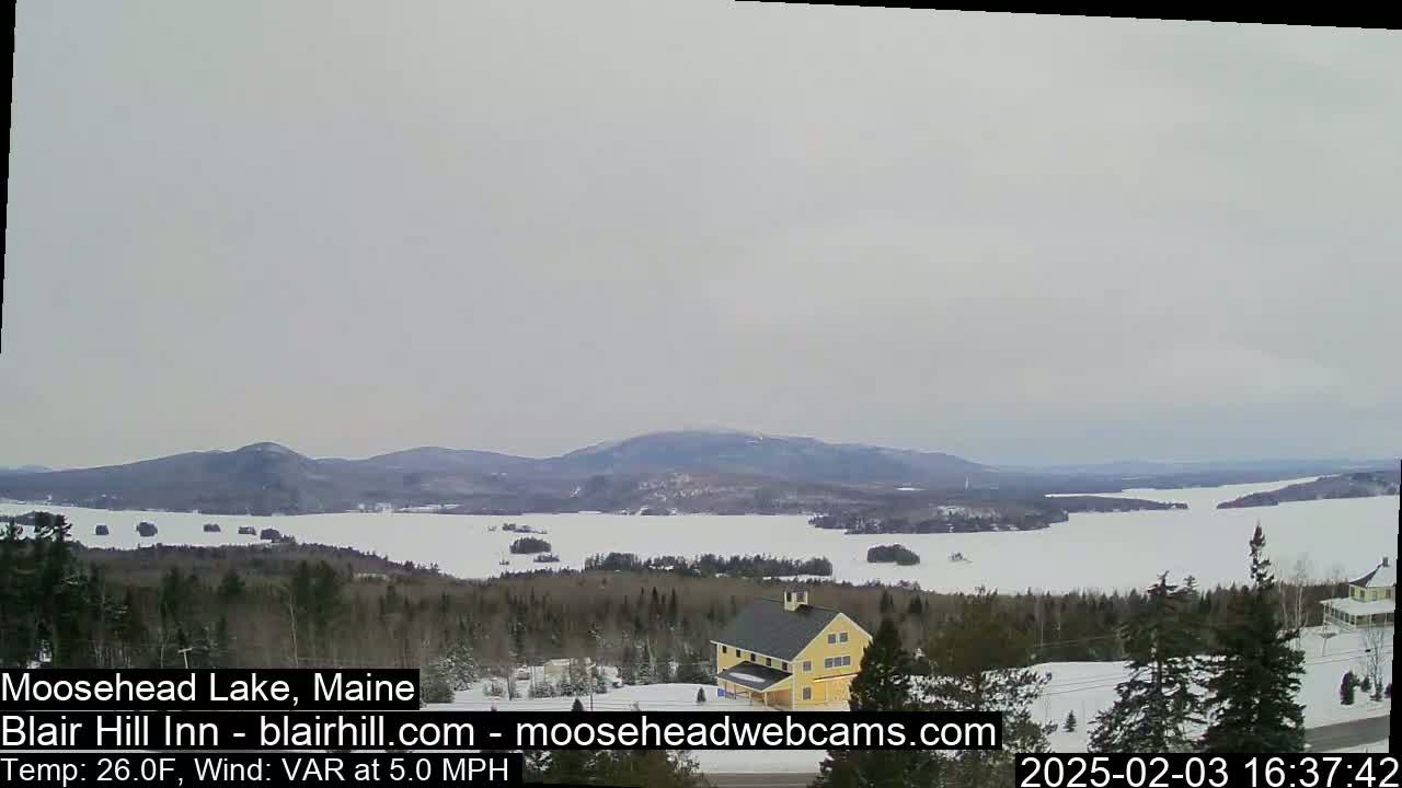 A partly frozen lake is seen from a snowy, wooded hillside, with a yellow building visible in the foreground and mountains in the distance under an overcast sky.