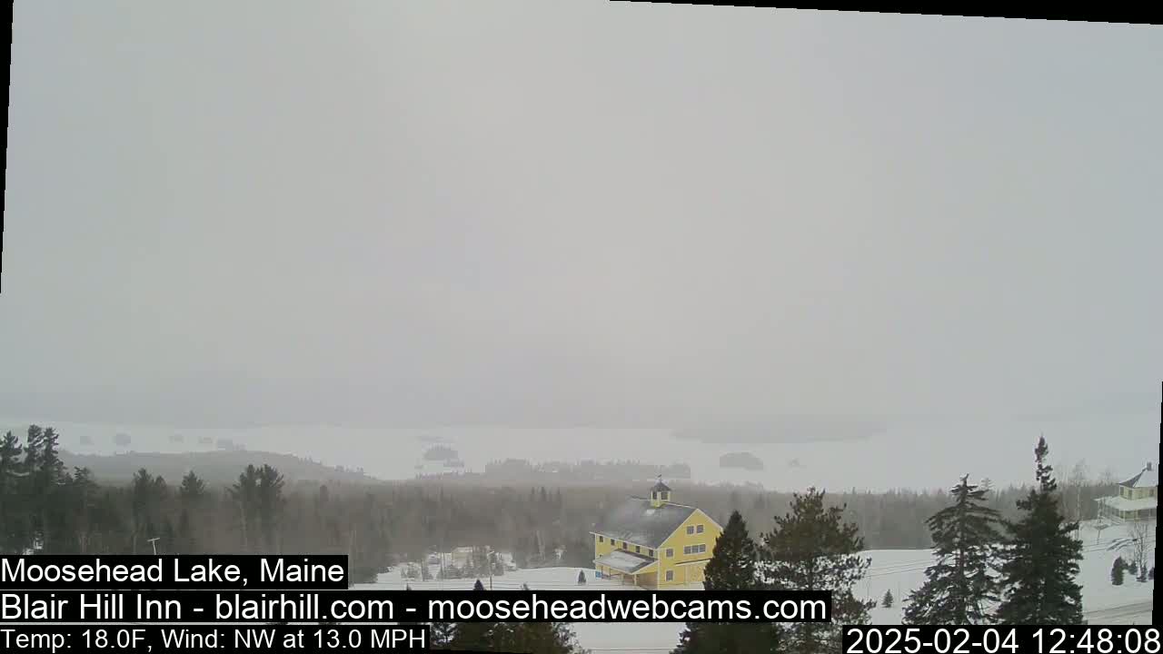 A snow-covered landscape with a yellow building and a lake in the distance under a hazy, overcast sky.