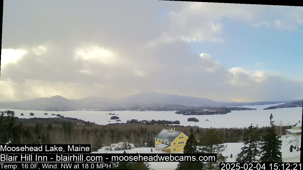 A partly cloudy, cold day shows a view of a snow-covered lake and distant mountains, with a yellow building in the foreground.