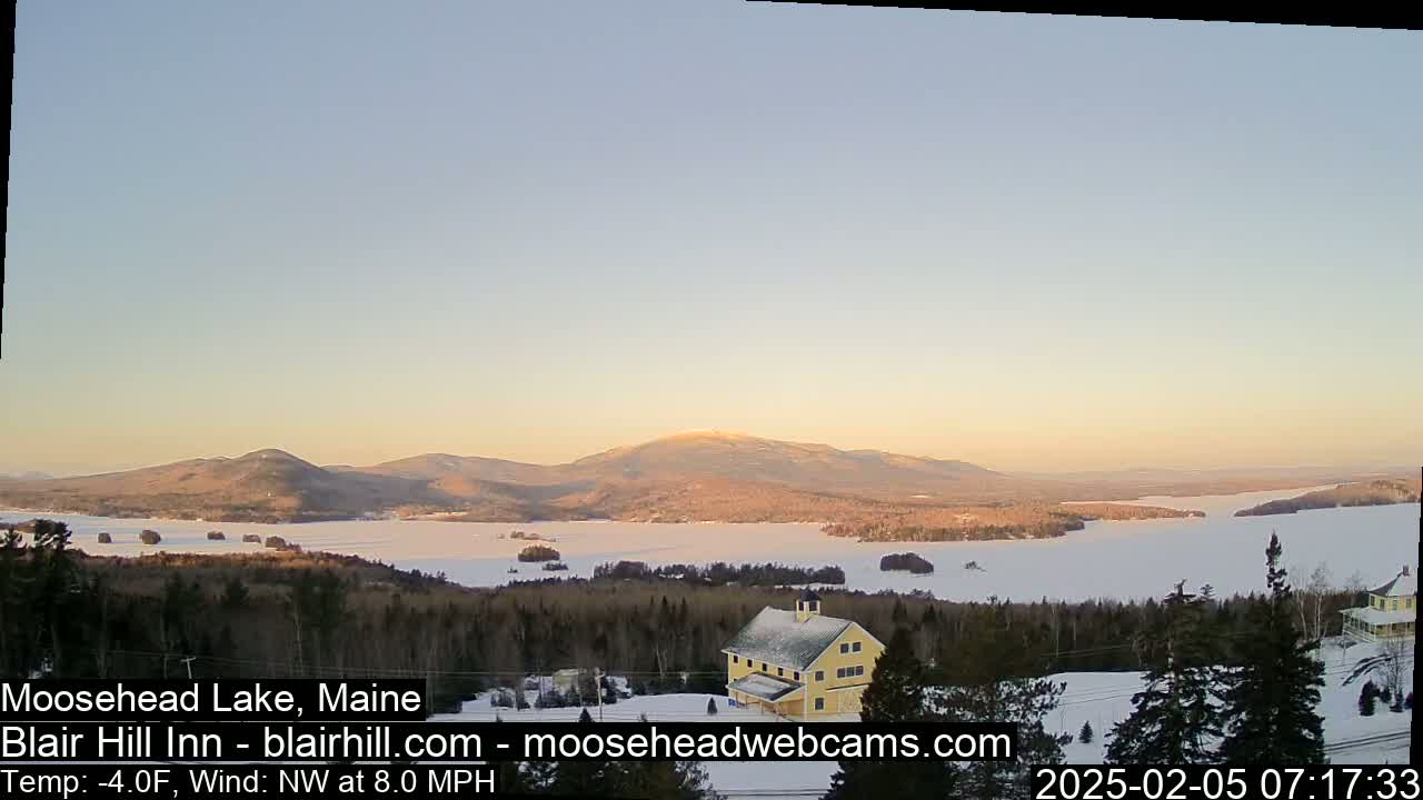 A frozen lake is surrounded by snow-covered hills and evergreen trees under a clear, light-blue sky at sunrise.
