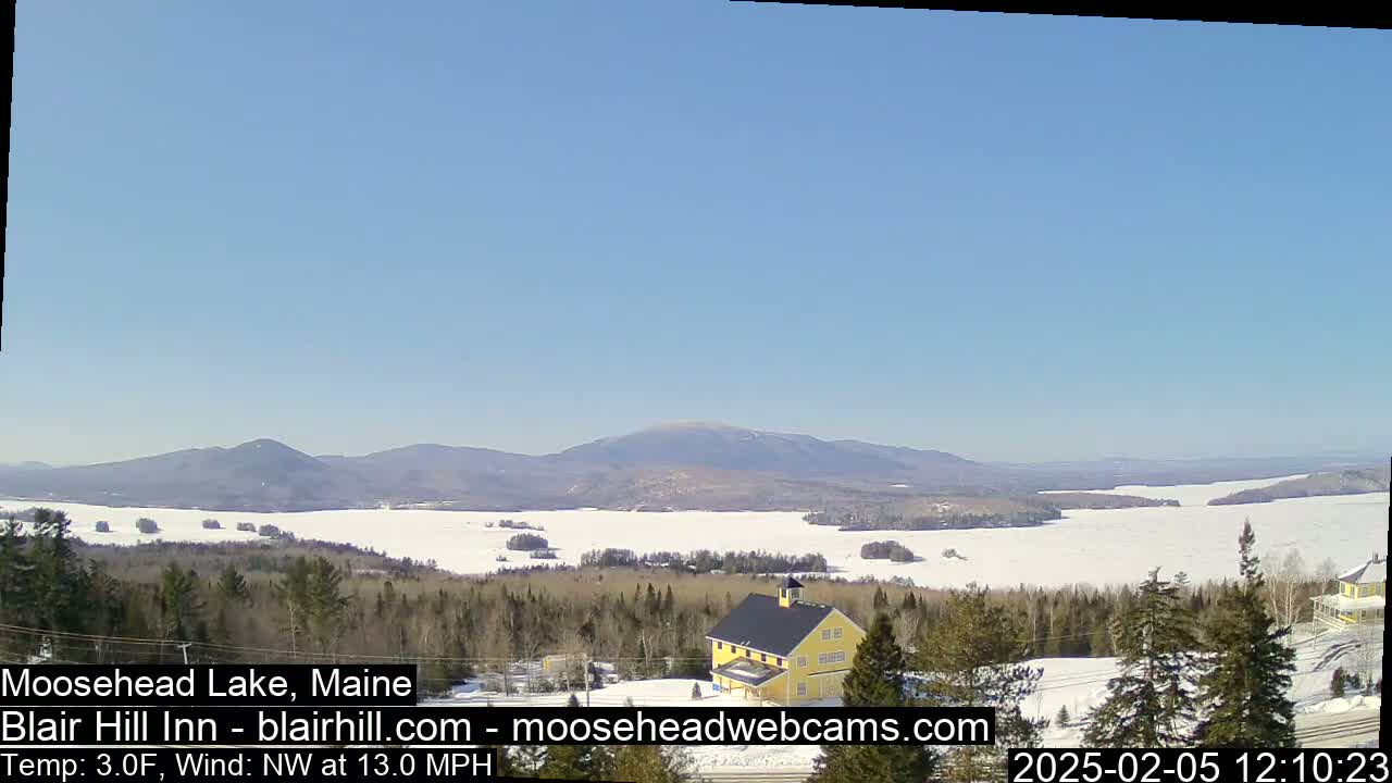 A mostly frozen lake stretches to snow-covered mountains under a clear blue sky, with a yellow building and evergreen trees in the foreground.