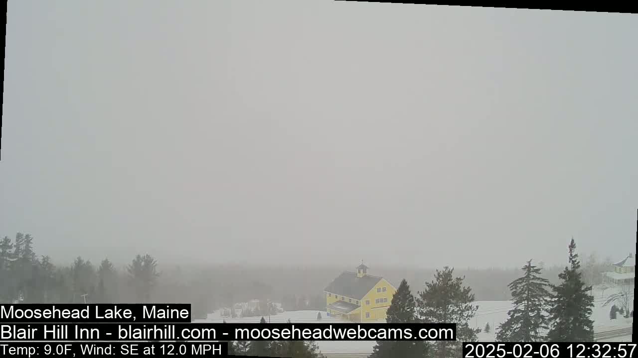 A hazy, snowy scene shows a yellow building among evergreens and a snow-covered landscape with a southeast wind at 12 mph and a temperature of 9°F.