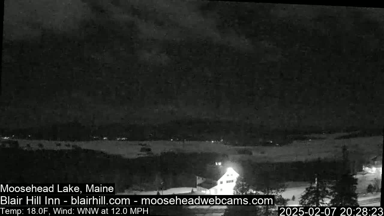 A nighttime, snowy landscape with a building lit up in the foreground under a cloudy sky, with a wind blowing from the west-northwest at 12 mph and a temperature of 18 degrees Fahrenheit.