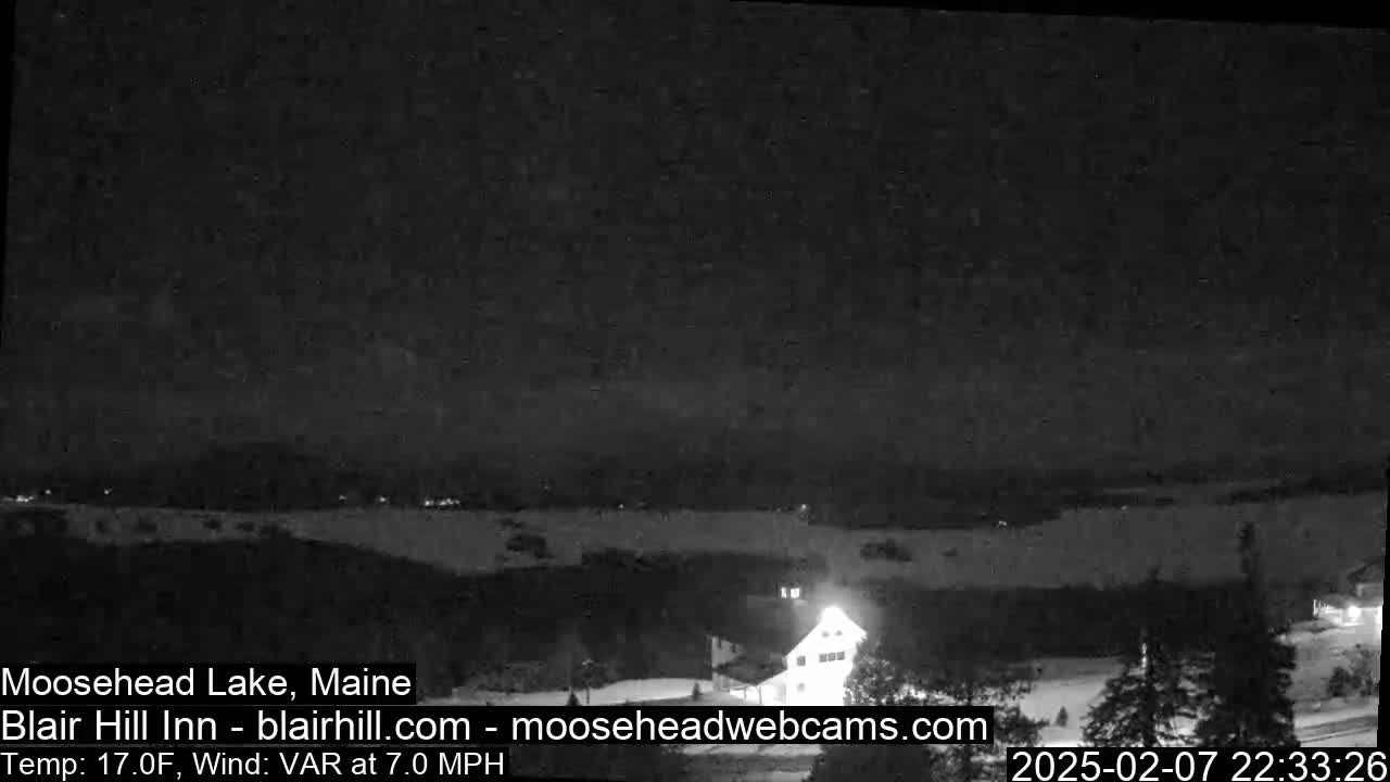 A nighttime, snow-covered landscape with a building illuminated, under a dark sky with a 17°F temperature and variable winds at 7.0 mph.