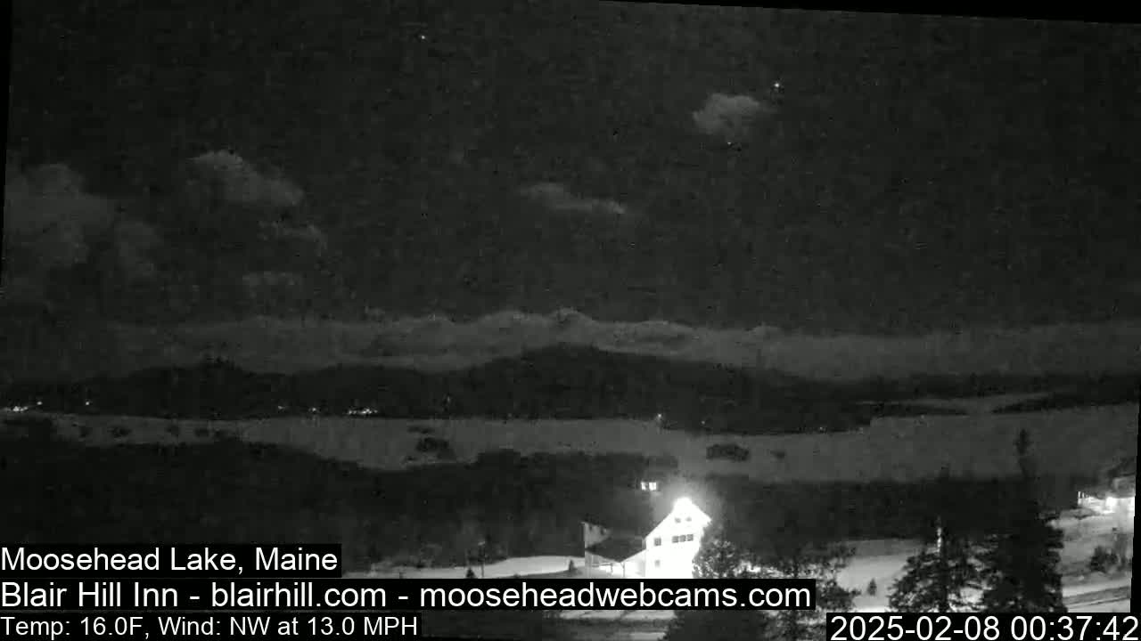 A nighttime view of a snow-covered landscape with a lake and a building under a partly cloudy sky, with a northwest wind at 13 mph and a temperature of 16°F.