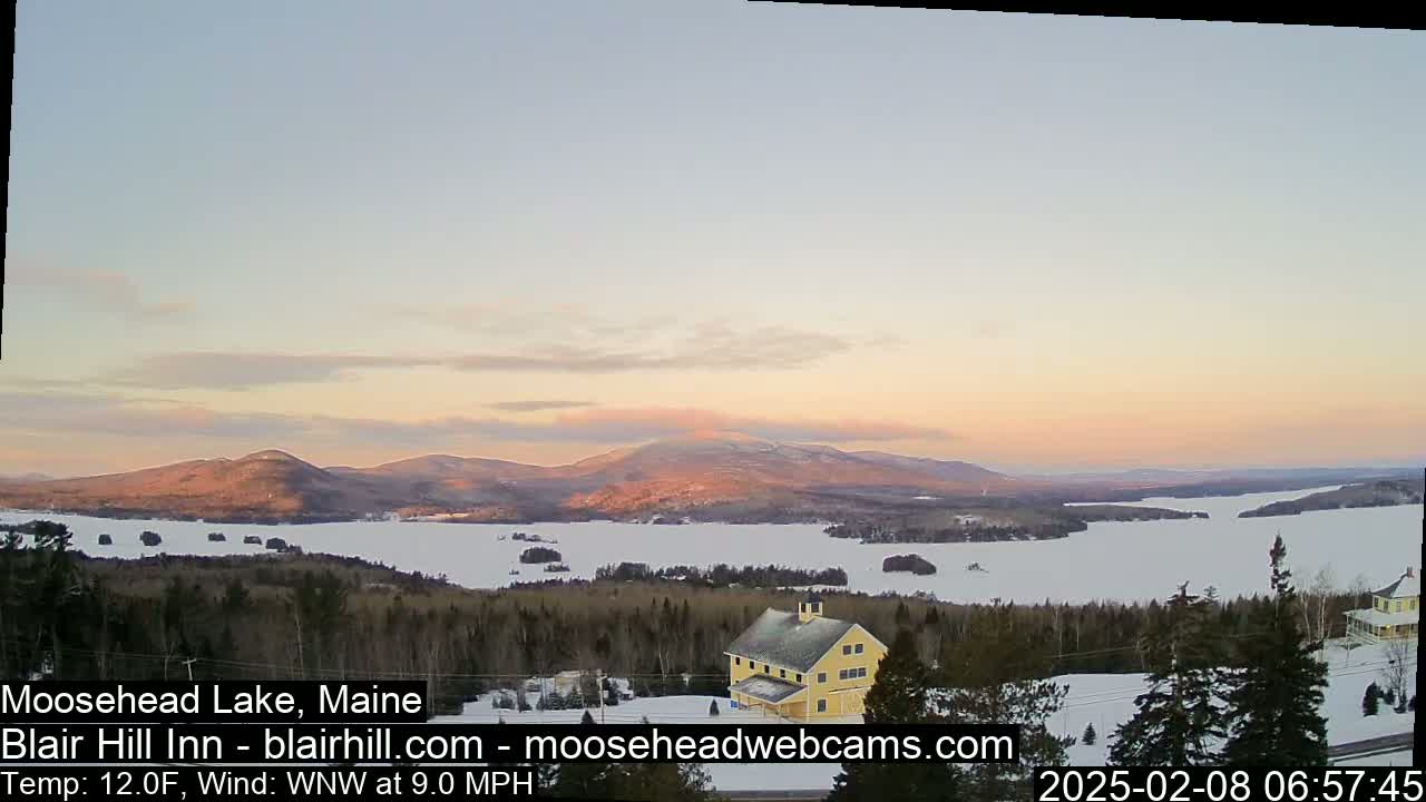 A snow-covered lake and surrounding forest are visible at sunset, with a yellow building in the foreground and mountains in the distance under a mostly clear sky.