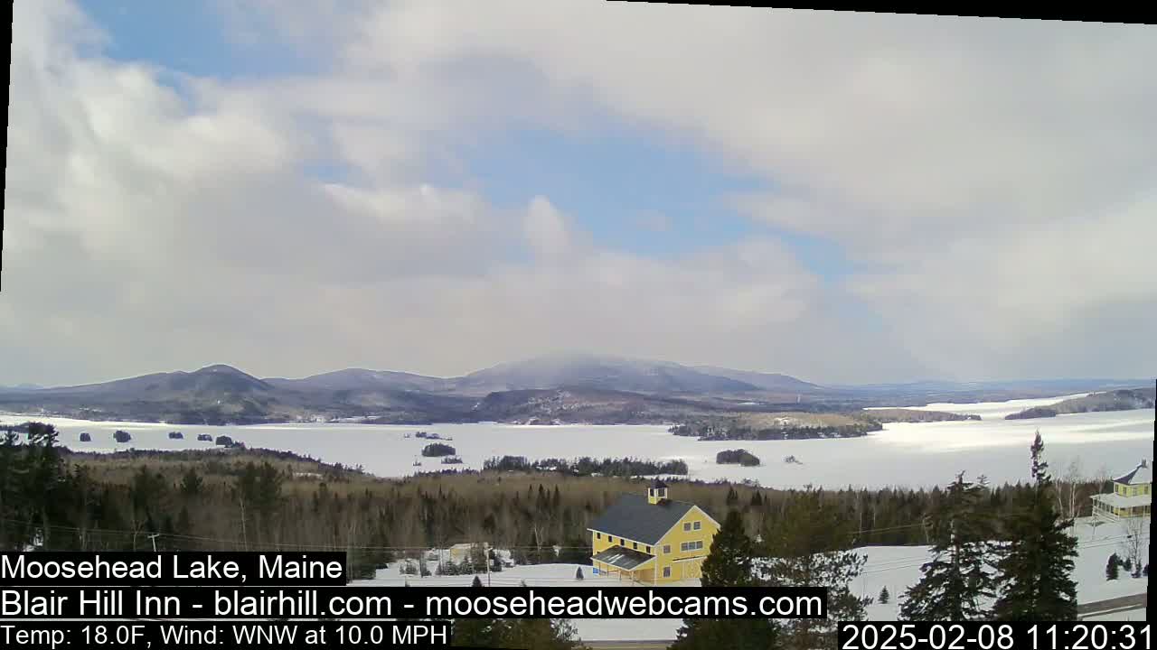 A partly cloudy day reveals a frozen lake nestled among snow-covered hills and evergreen trees, with a yellow building visible in the foreground.