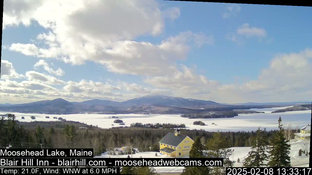 A partly cloudy day reveals a frozen lake nestled among snow-covered hills and evergreen trees, with a yellow building visible in the foreground.