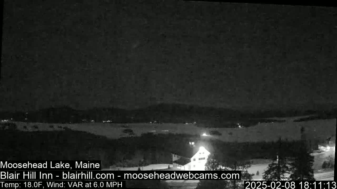 A nighttime view of a snow-covered landscape near a lake under a dark sky with a variable wind at 6 mph and a temperature of 18°F.