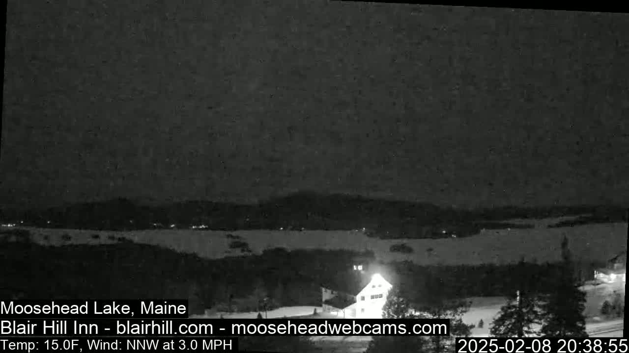 A nighttime, snow-covered landscape shows a lake with a building and trees illuminated by lights under a dark sky with a 15°F temperature and a 3 mph northwest wind.