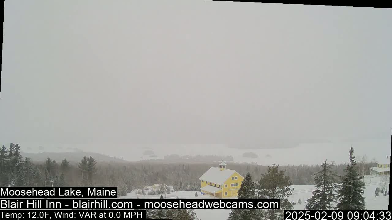 A snow-covered landscape shows a yellow barn, evergreen trees, and a lake in the distance under a hazy, overcast sky.