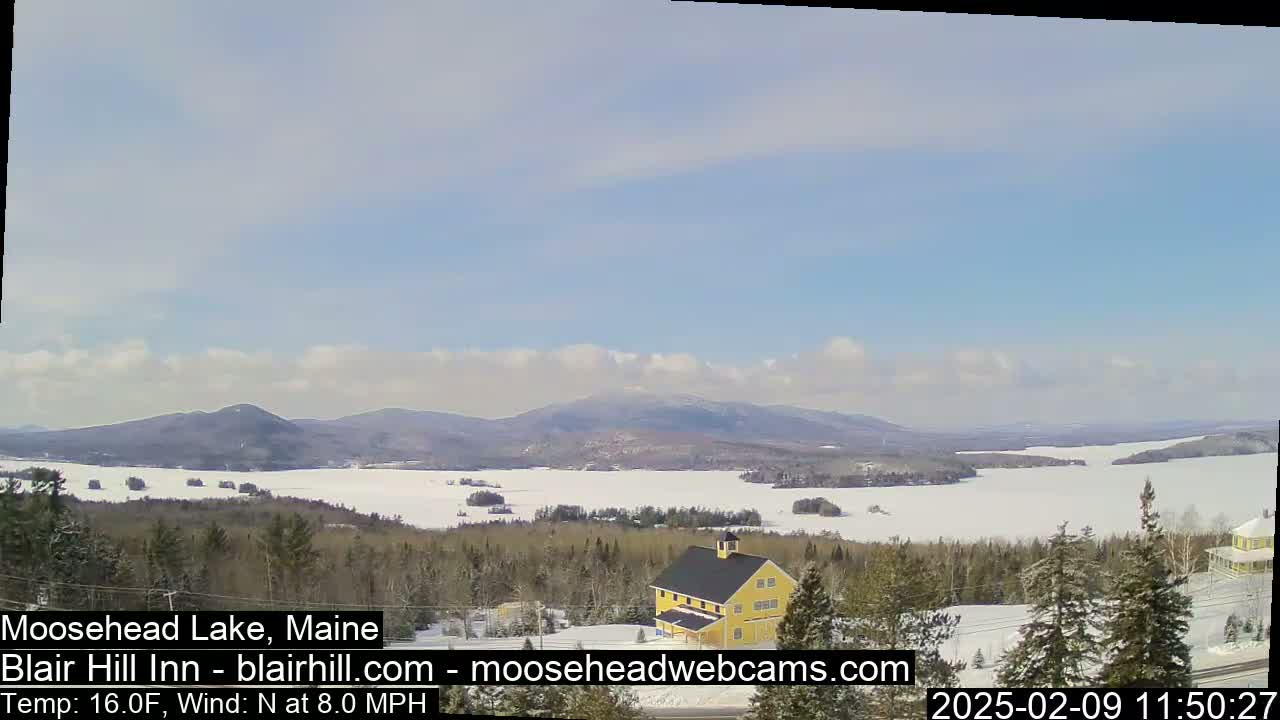 A partly cloudy, cold day shows a snow-covered lake and mountains in the distance, with a yellow building in the foreground.