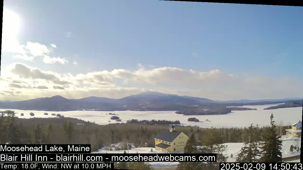 A partly cloudy, cold day with a northwest wind reveals a snow-covered lake and distant mountains, viewed from a high vantage point overlooking a yellow building and a snow-covered landscape.