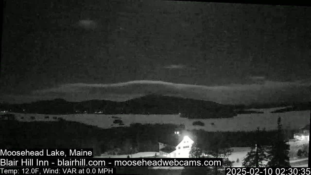 A nighttime, snow-covered landscape shows a lake and distant hills under a dark sky with a thin layer of clouds, and a building with lights is visible in the foreground.