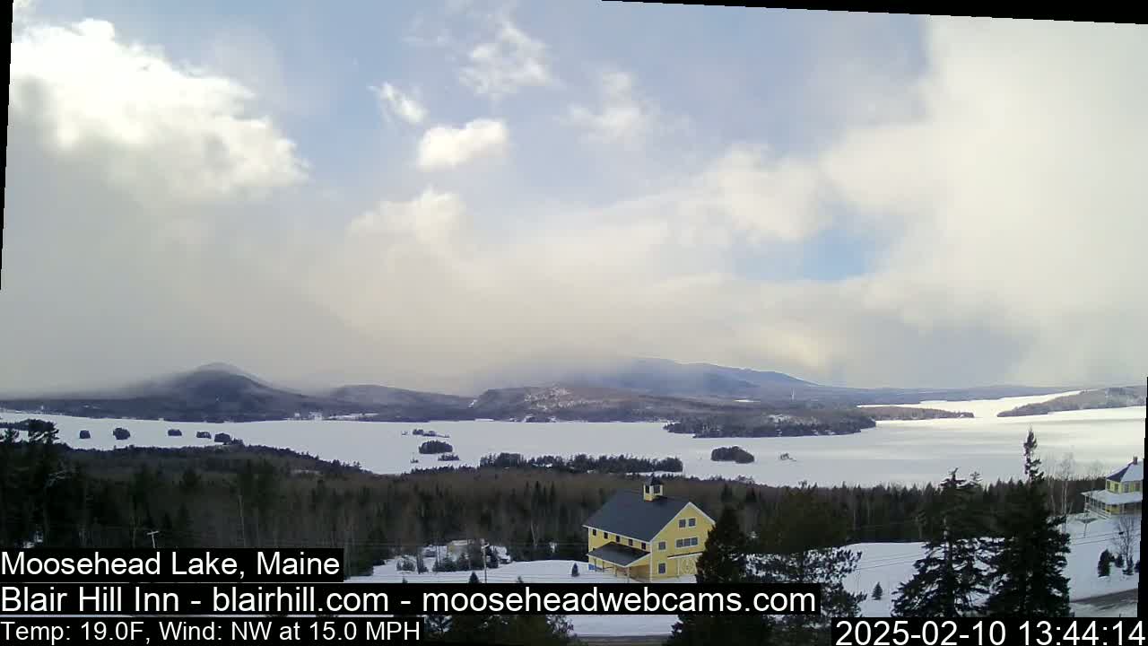 A partly cloudy, cold day with a northwest wind reveals a snow-covered lake and distant mountains, observed from a vantage point near a yellow building.
