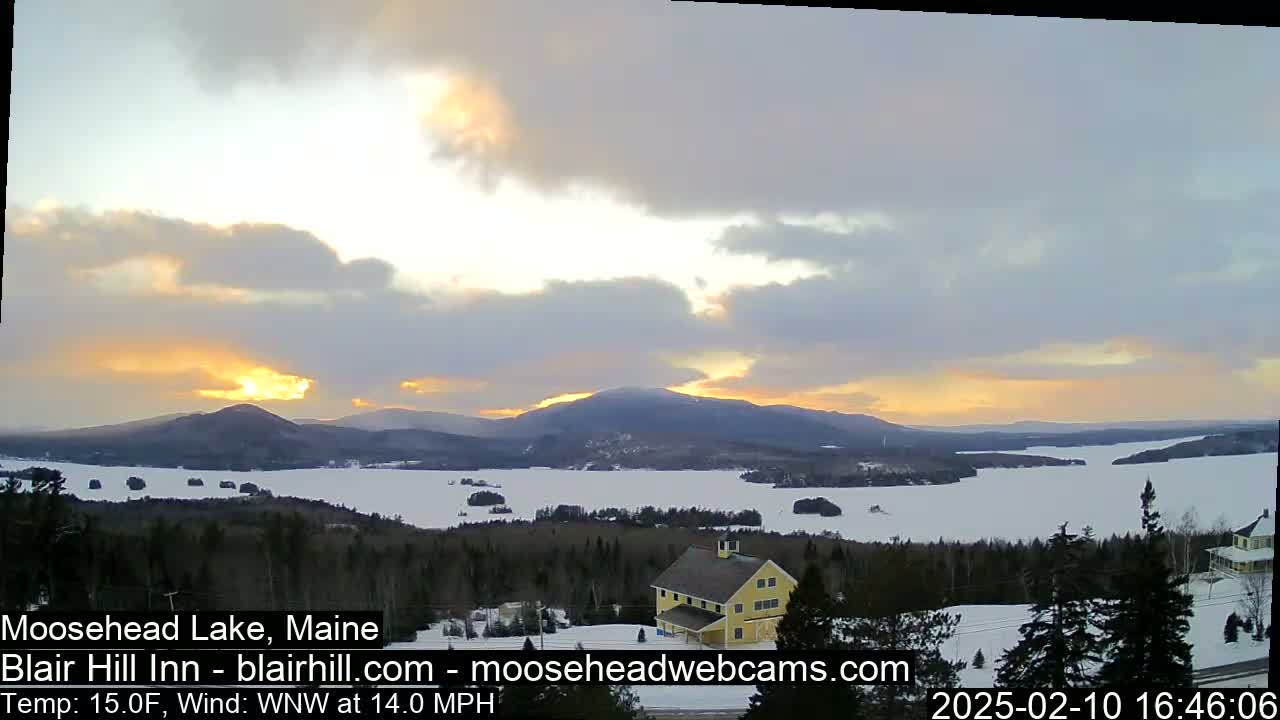 A snow-covered landscape overlooks a partially frozen lake at sunset, with mountains in the background and a yellow building in the foreground under partly cloudy skies and a 14 mph wind.