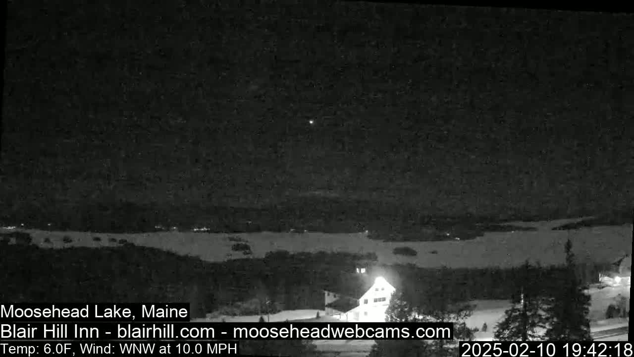 A nighttime, snowy landscape shows a body of water under a dark sky with a single, bright light visible in the distance.
