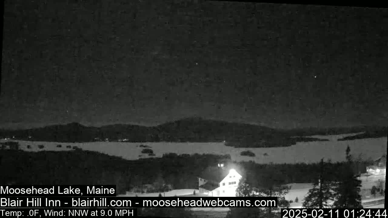 A nighttime view of a snow-covered landscape shows a lake and distant hills under a clear, starry sky, with a building lit up in the foreground, and a 9.0 mph wind from the north-northwest at 0°F.