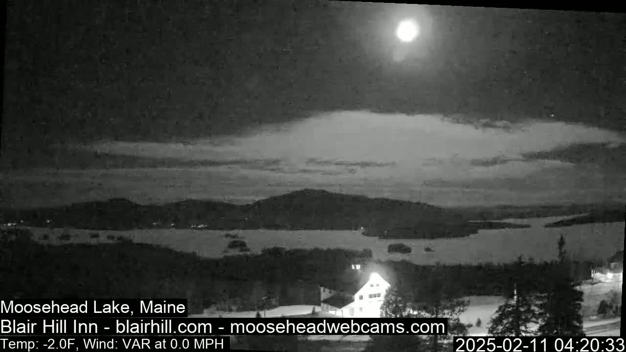 A nighttime view of a lake and snowy landscape under a bright moon and partly cloudy sky.