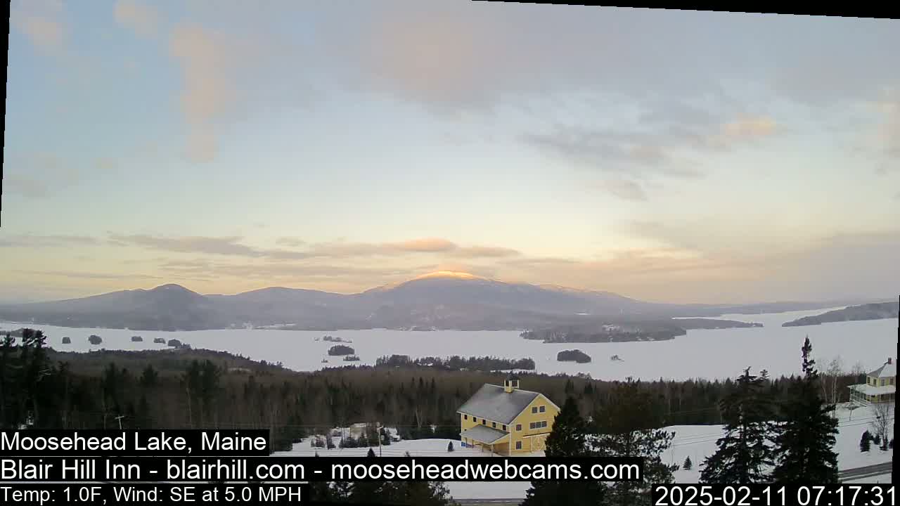 A snow-covered landscape reveals a frozen lake surrounded by dark evergreen trees, distant mountains, and a yellow building at sunset under a partly cloudy sky.