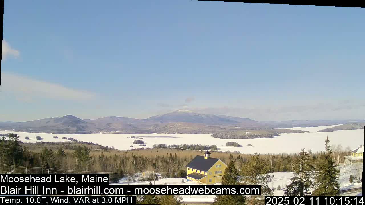A snow-covered lake and distant mountains are visible under a clear blue sky, with a yellow building in the foreground and a light wind.