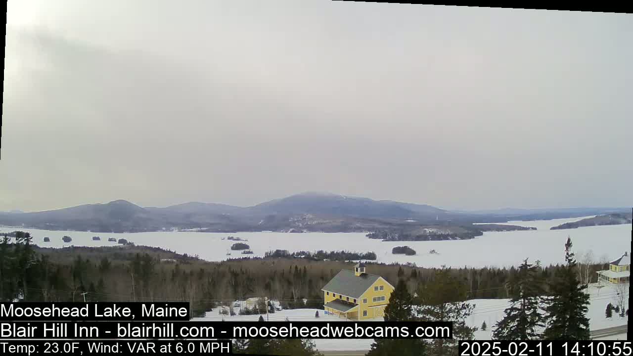 A snow-covered lake with distant mountains is viewed from a wooded hillside, under an overcast sky; a yellow building is visible in the foreground.