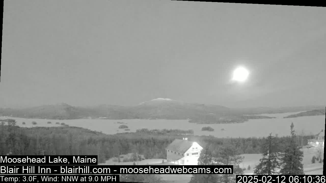A nighttime, snow-covered landscape shows a lake, distant hills, and a building, under a bright, possibly moonlit sky with a 3°F temperature and a 9 mph northwest wind.