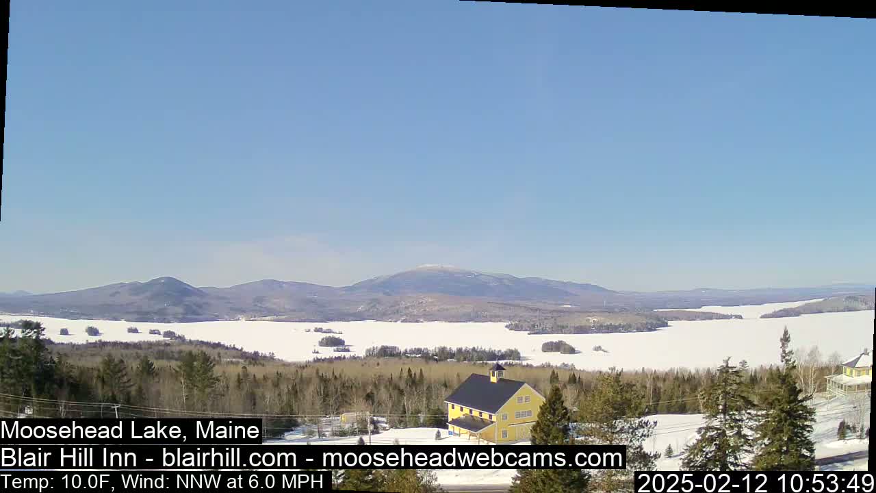 A snow-covered lake and distant mountains are visible under a clear blue sky, with a yellow building in the foreground and a light north-northwest wind at 6 mph and a temperature of 10 degrees Fahrenheit.