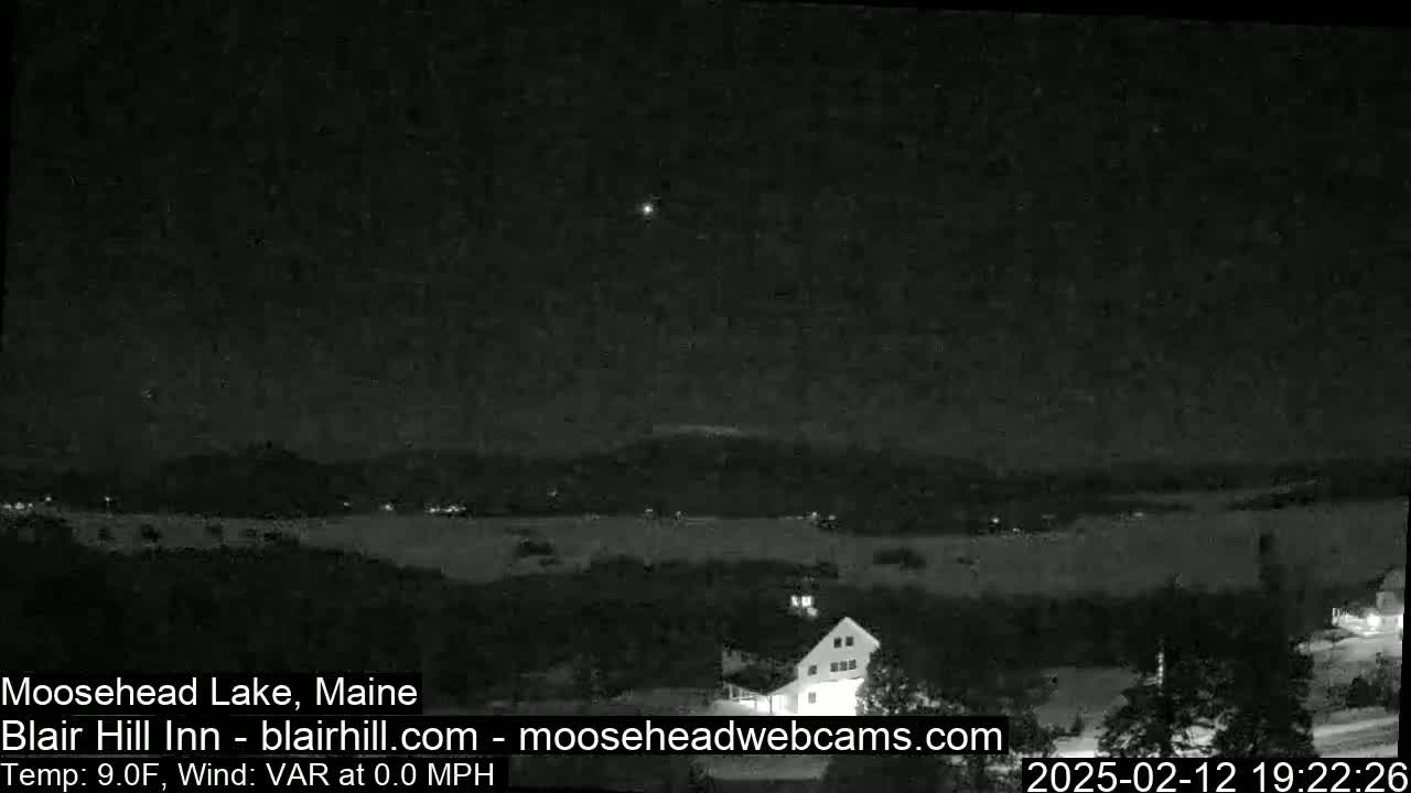 A nighttime, low-light view of a lake and a building, under a clear sky with a single bright light visible in the distance.