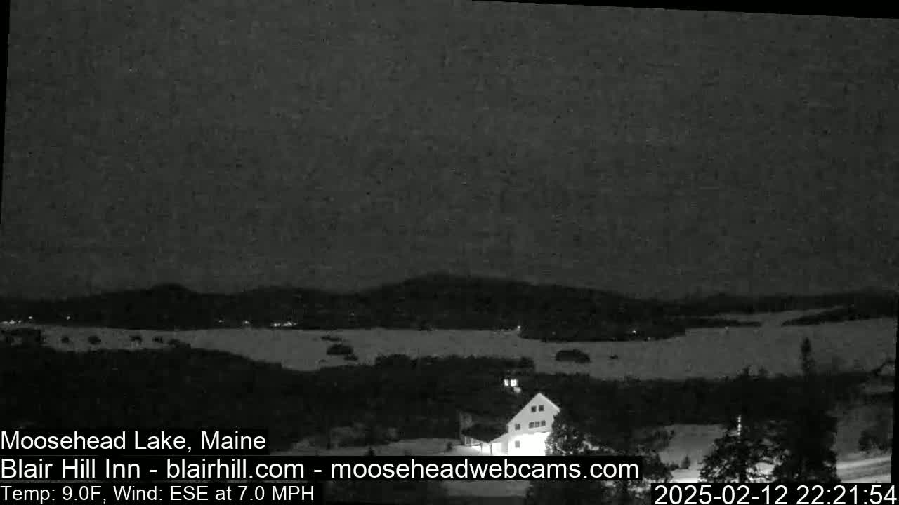 A nighttime view of a snow-covered landscape with a lake and a building, under a dark sky with a 9°F temperature and a 7 mph east-southeast wind.