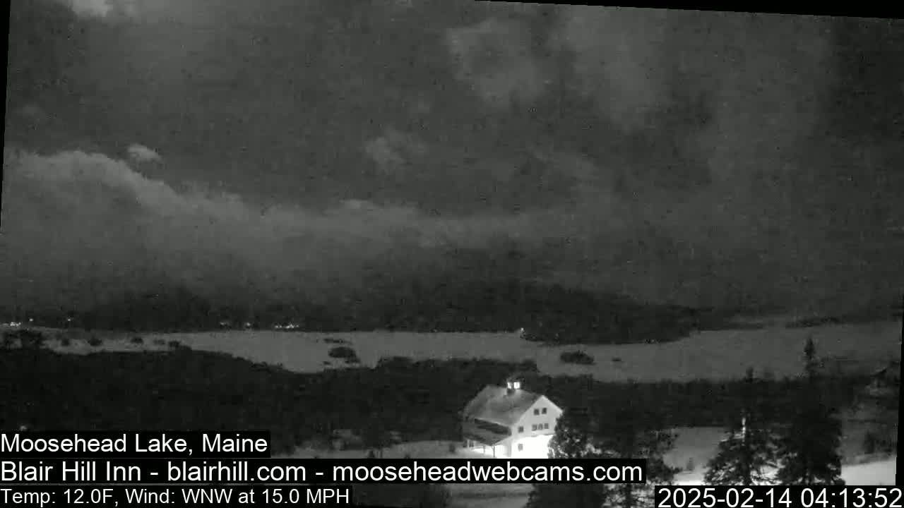 A nighttime, snowy landscape shows a body of water with a building and trees under a cloudy sky with a 12°F temperature and 15 mph wind.