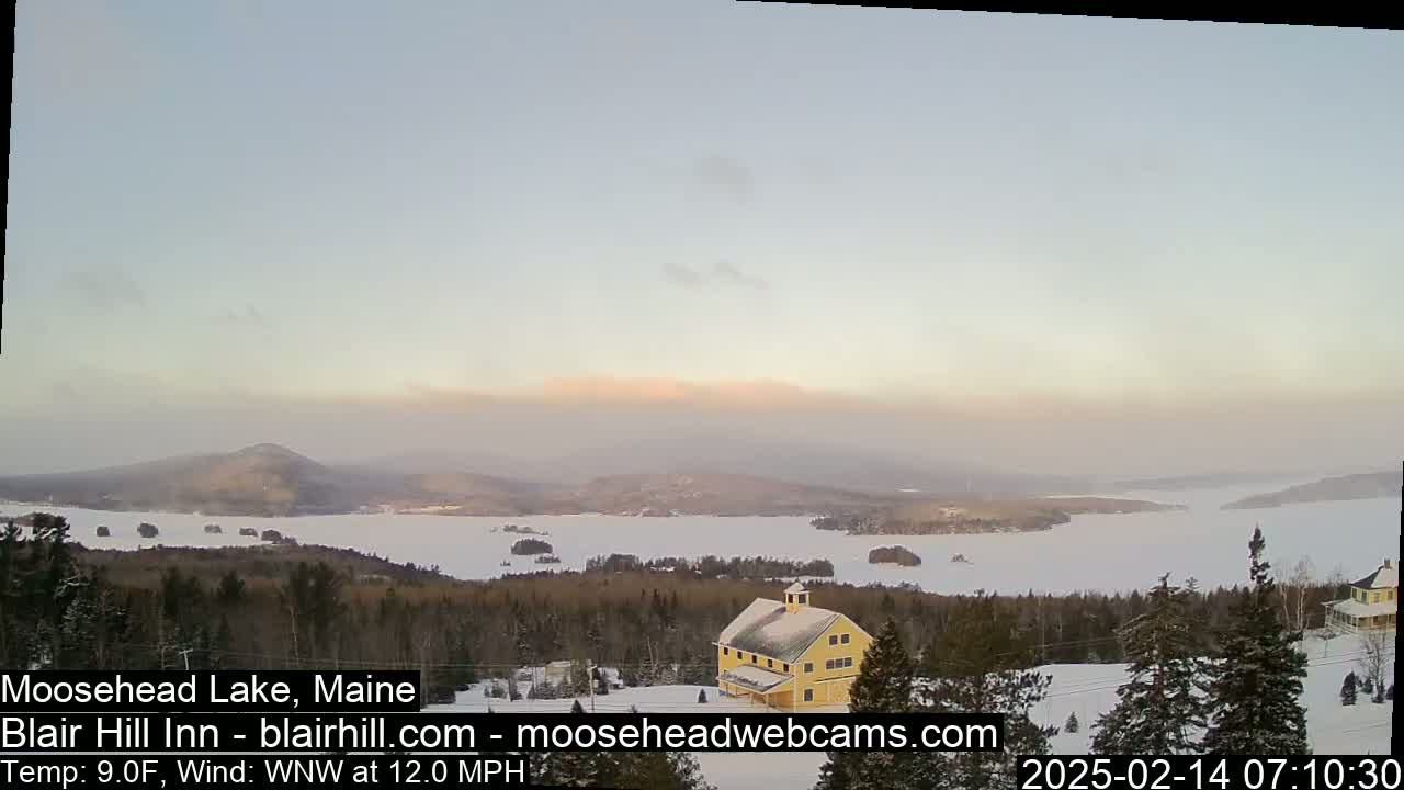 A yellow building sits on a snow-covered hill overlooking a frozen lake and distant mountains under a hazy, light-pink sunrise.