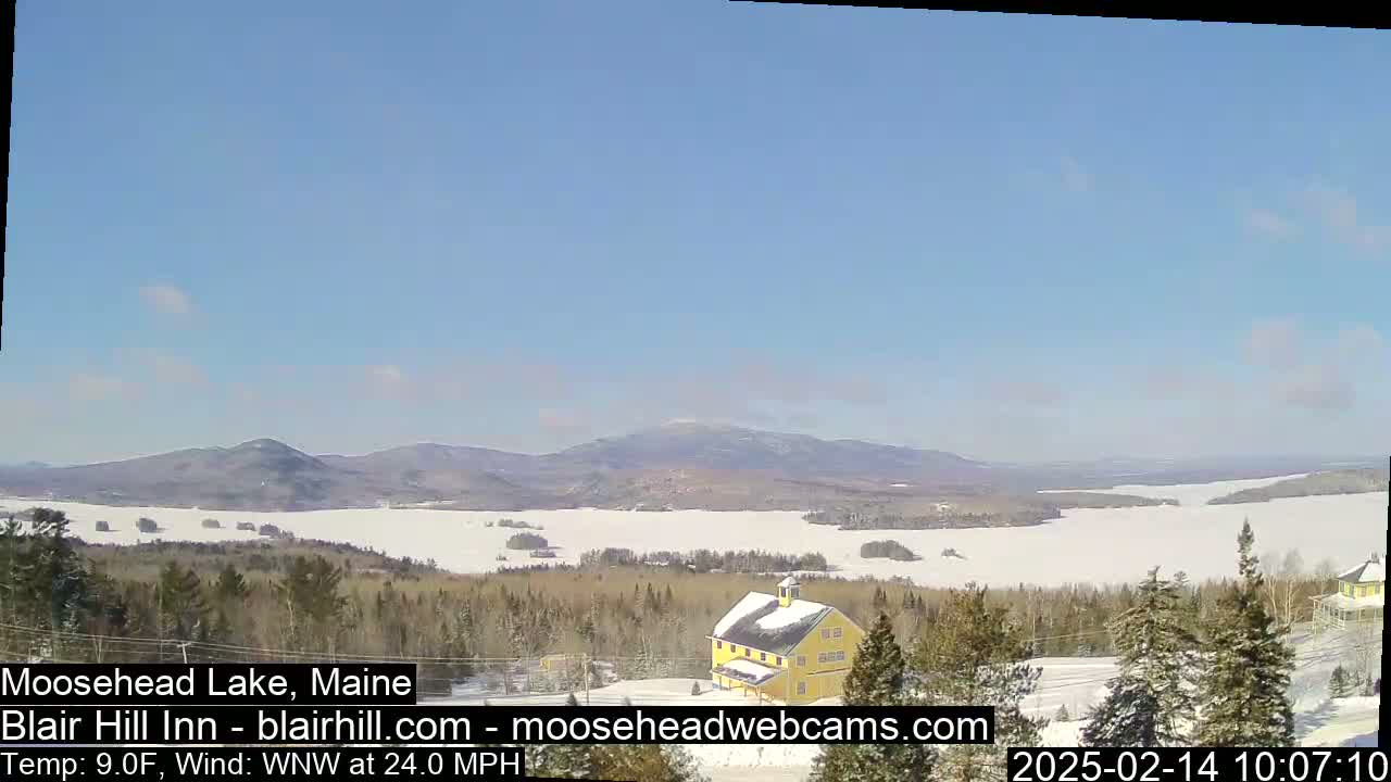 A yellow building sits on a snow-covered hill overlooking a frozen lake and distant mountains under a clear blue sky with a WNW wind at 24.0 mph.