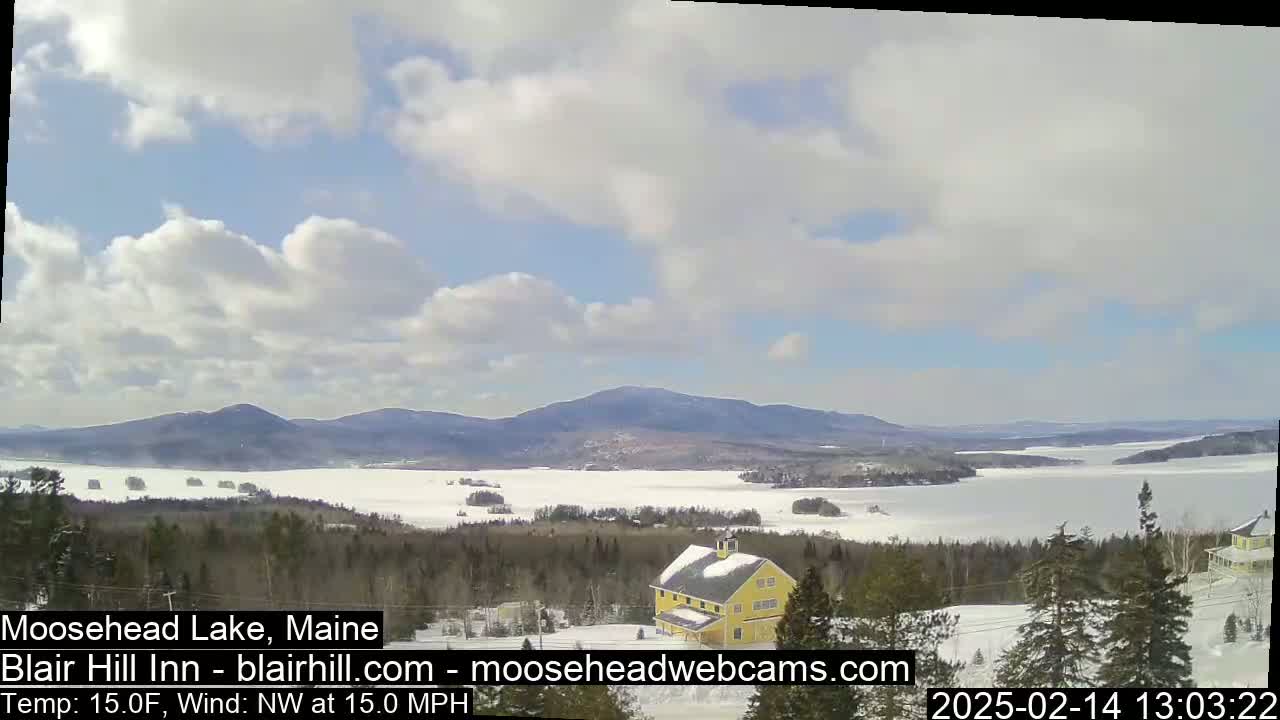 A partly cloudy, cold day with a northwest wind displays a view of a frozen lake, a yellow building on a snow-covered hillside, and distant mountains.