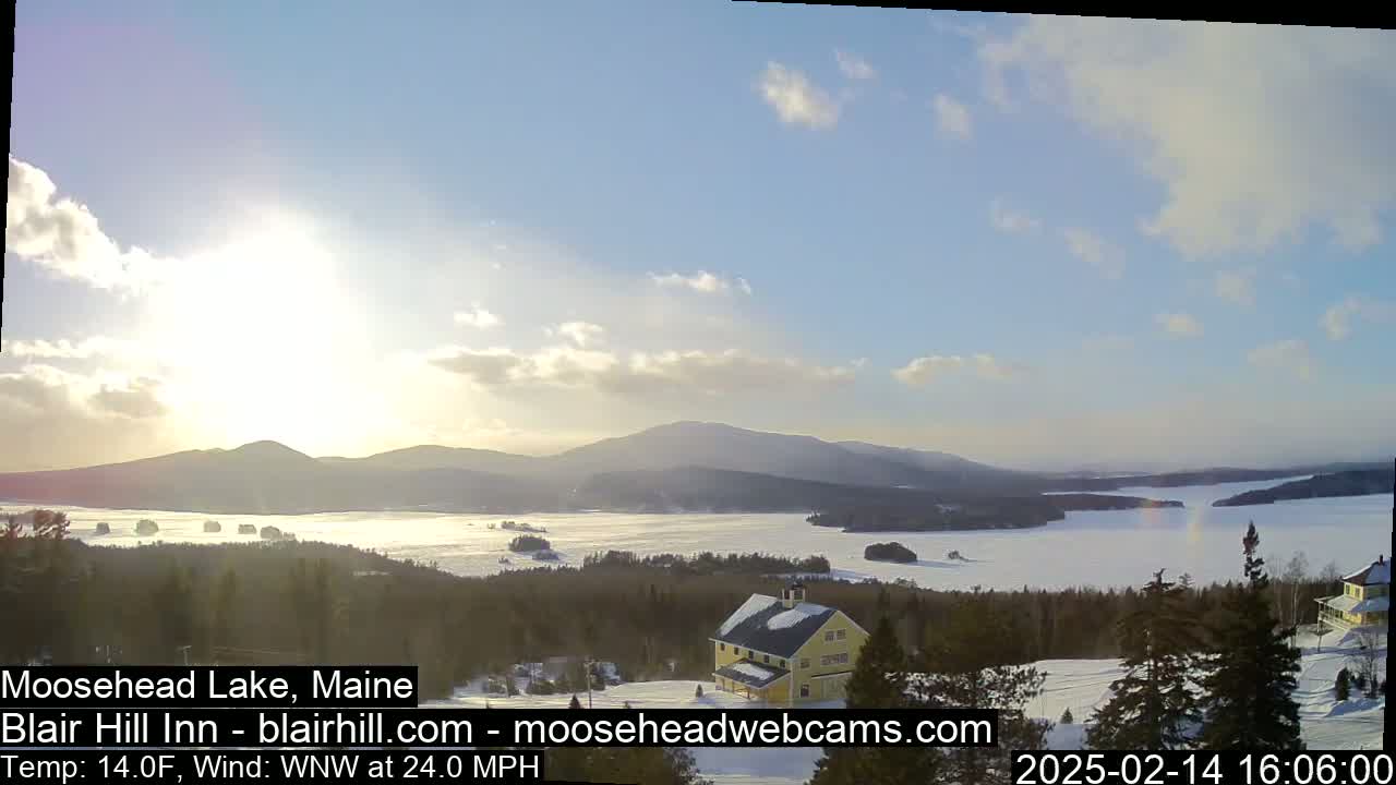 A mostly sunny, cold day with a strong wind shows a snow-covered landscape featuring a frozen lake, distant mountains, and a yellow building.