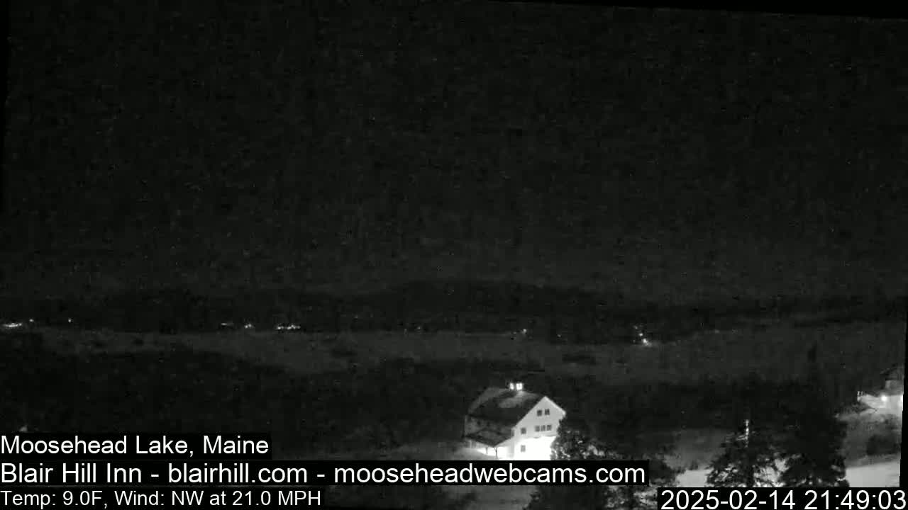 A nighttime, low-light image shows a snow-covered landscape with a building and a body of water under a dark sky with a northwest wind at 21 mph and a temperature of 9°F.