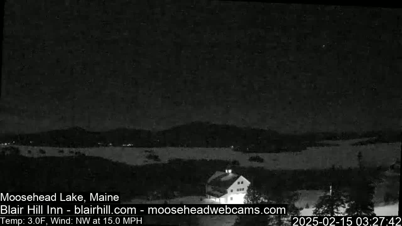 A nighttime view of a snowy landscape with a building and distant mountains under a clear, dark sky with a northwest wind.