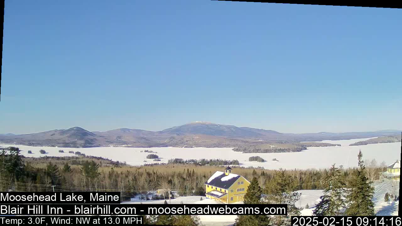 A yellow building sits on a snow-covered hill overlooking a frozen lake and distant mountains under a clear blue sky.
