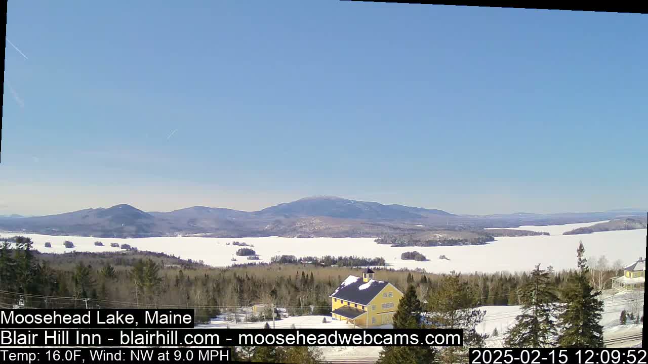 A partly frozen lake is nestled among snow-covered hills and evergreen trees under a clear blue sky.