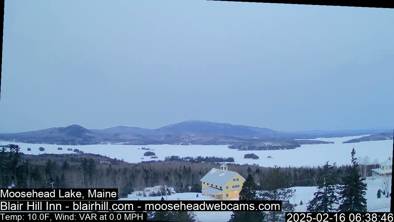 A snow-covered landscape featuring a frozen lake, distant mountains, and a yellow building nestled among evergreen trees under a pale, overcast sky.