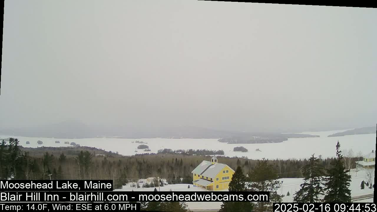 A hazy, snowy landscape shows a partially frozen lake, a yellow barn, and a snow-covered forest under a gray sky.